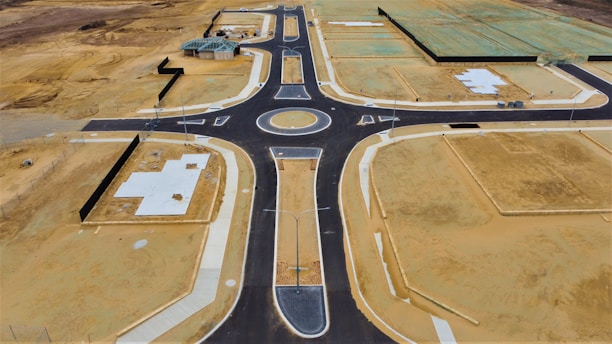 Aerial view of a newly constructed road with a roundabout in a development area. The site features patches of bare soil, concrete foundations, and partially constructed buildings. The surrounding land appears undeveloped, with empty plots of dirt and some green-covered sections indicating future construction.