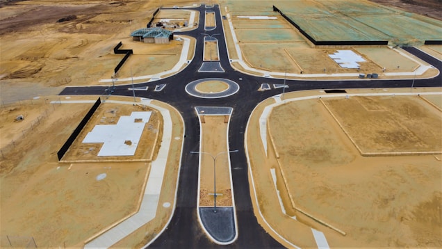 Aerial view of a newly constructed road with a roundabout in a development area. The site features patches of bare soil, concrete foundations, and partially constructed buildings. The surrounding land appears undeveloped, with empty plots of dirt and some green-covered sections indicating future construction.