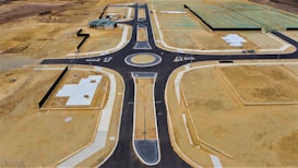 Aerial view of a newly constructed road with a roundabout in a development area. The site features patches of bare soil, concrete foundations, and partially constructed buildings. The surrounding land appears undeveloped, with empty plots of dirt and some green-covered sections indicating future construction.