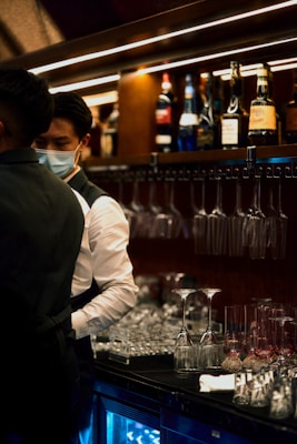 A bartender wearing a mask is preparing drinks behind a bar counter, which is lined with rows of empty wine and champagne glasses. The background features various bottles of alcohol on shelves, creating a dimly lit and cozy atmosphere.