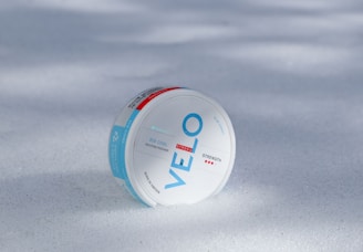A round white container labeled 'VELO' with blue and red accents lies on a surface covered in snow. The product is described as 'ICE COOL NICOTINE POUCHES' with 'STRONG' strength indicators. The words 'BORN IN SWEDEN' are also visible on the package.