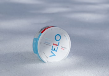 A round white container labeled 'VELO' with blue and red accents lies on a surface covered in snow. The product is described as 'ICE COOL NICOTINE POUCHES' with 'STRONG' strength indicators. The words 'BORN IN SWEDEN' are also visible on the package.