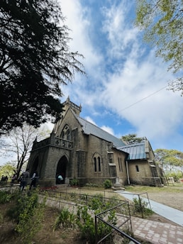 A historic stone church stands prominently with a tall, steeply pitched roof and Gothic-style architecture. The sky is bright with a few clouds, and trees with green leaves frame the scene. Several people are near the entrance, which features pointed arch doorways. The surrounding garden includes neatly maintained plants and a paved walkway.