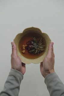 Two hands holding a beige, scalloped bowl filled with a brown liquid and various herbal ingredients, such as leaves and berries.