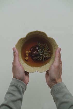 Two hands holding a beige, scalloped bowl filled with a brown liquid and various herbal ingredients, such as leaves and berries.