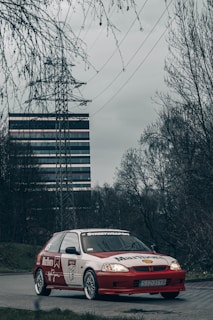 A modified red and white car with advertising decals is parked on a road in an urban or industrial area. The surroundings are marked by bare trees and a tall building in the background, along with power lines and a pylon adding to the industrial setting. The sky appears overcast, contributing to a moody ambiance.