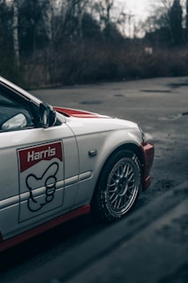 The image features a close-up of a white car with alloy wheels. The car has a prominent logo and text 'Harris' on the side, with a red and black design. It is parked on a paved area surrounded by bare trees, suggesting a cold, possibly winter, setting.