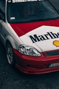 A customized car features a distinctive red and white paint job with large Marlboro and Shell logos. The car is equipped with aftermarket components, including sporty rims and a front bumper. The windshield displays a 'Streetweapon' decal, and there is evident branding inside the car on the seats.