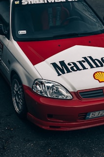 A customized car features a distinctive red and white paint job with large Marlboro and Shell logos. The car is equipped with aftermarket components, including sporty rims and a front bumper. The windshield displays a 'Streetweapon' decal, and there is evident branding inside the car on the seats.