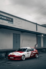 A racing car with a red and white livery is parked in front of an industrial building. The car features various sponsor logos, including Honda, Marlboro, and Shell. The building in the background has a metal exterior with closed doors and an industrial sign at the top.