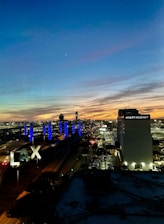 A vibrant cityscape during sunset, featuring a clear sky with gradients of blue, orange, and purple. Prominent in the foreground are large illuminated 'LAX' letters beside several roads with moving vehicles. In the background, tall blue lights create a striking visual, and a Hyatt Regency hotel is visible alongside a busy airport environment.