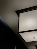 A modern ceiling light fixture with geometric shapes and a minimalistic design. The image shows a large square light panel with a small cylindrical bulb in the center, set against a dark ceiling with contrasting shades.