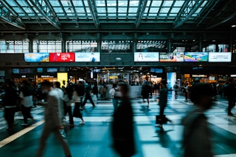 A bustling train station with many people walking in different directions. The high ceiling with glass panels allows natural light to fill the space. Large digital advertisement screens line the upper walls, displaying various colorful ads and information. The floor has a reflective finish, creating a dynamic interplay of light and shadow.