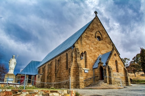 A stone church with pointed roofs and a large entrance stands under a cloudy sky. A statue of a religious figure holding a staff is visible to the side. The church is surrounded by a simple stone fence and is elevated slightly with steps leading up to the entrance.