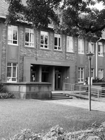 A brick building with a sign that reads 'Teologie Theology' is situated in a serene environment. The building has large windows and a flat roof. In front, there is a grassy area and a pathway leading to the entrance. A streetlamp stands near the path, and trees with dense foliage partially shade the building.