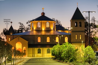 A welcoming warm light shining from a church window surrounded by trees at dusk.