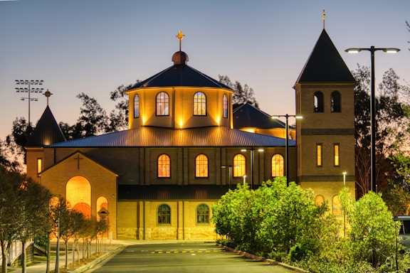 A welcoming warm light shining from a church window surrounded by trees at dusk.