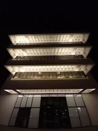 A modern building with a unique layered design, featuring large glass windows and illuminated sections that stand out against the night sky. The building has multiple levels with a symmetrical structure and a sleek, contemporary appearance.