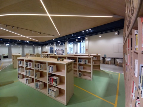 A modern library interior with wooden bookshelves filled with books. The ceiling features geometric patterns with integrated lighting. The floor is green with yellow markings. In the background, tables and chairs are arranged for studying or reading.