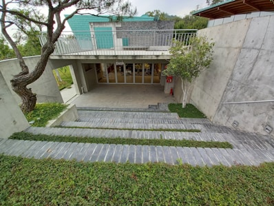 A modern architectural structure with concrete walls, an overhang, and a green roof. There are steps leading down to an entrance with glass doors labeled 'shop.' The surrounding landscape includes neatly trimmed grass and trees, providing a harmonious blend of nature and structure.