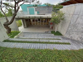 A modern architectural structure with concrete walls, an overhang, and a green roof. There are steps leading down to an entrance with glass doors labeled 'shop.' The surrounding landscape includes neatly trimmed grass and trees, providing a harmonious blend of nature and structure.