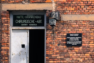 A brick wall features two signs: one above a metal door with German text indicating a forbidden entry, and another to the right detailing a national exhibition about the persecution and deportation of Jews in the Netherlands during 1940-1945. A lantern is mounted on the wall displaying the number 21.