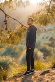 A person stands on a sunlit path surrounded by nature, gazing upwards with a thoughtful expression. The golden hour light filters through the branches of a nearby tree, casting a warm glow over the scene. The person is wearing a dark jacket and black pants, with hands in the pockets.