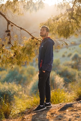 A person stands on a sunlit path surrounded by nature, gazing upwards with a thoughtful expression. The golden hour light filters through the branches of a nearby tree, casting a warm glow over the scene. The person is wearing a dark jacket and black pants, with hands in the pockets.