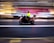 A motorcyclist wearing a bright yellow jacket rides a red motorcycle through a city street. The background is blurred, suggesting motion and speed, with lights and buildings creating streaks of color.