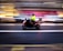 A motorcyclist wearing a bright yellow jacket rides a red motorcycle through a city street. The background is blurred, suggesting motion and speed, with lights and buildings creating streaks of color.