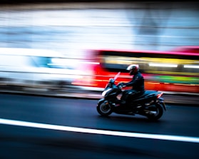 A motorcyclist wearing a helmet is riding at speed on a dark road. Motion blur suggests movement, with bright streaks of light in the background indicating passing vehicles, creating a dynamic and fast-paced scene.