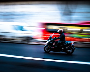 A motorcyclist wearing a helmet is riding at speed on a dark road. Motion blur suggests movement, with bright streaks of light in the background indicating passing vehicles, creating a dynamic and fast-paced scene.