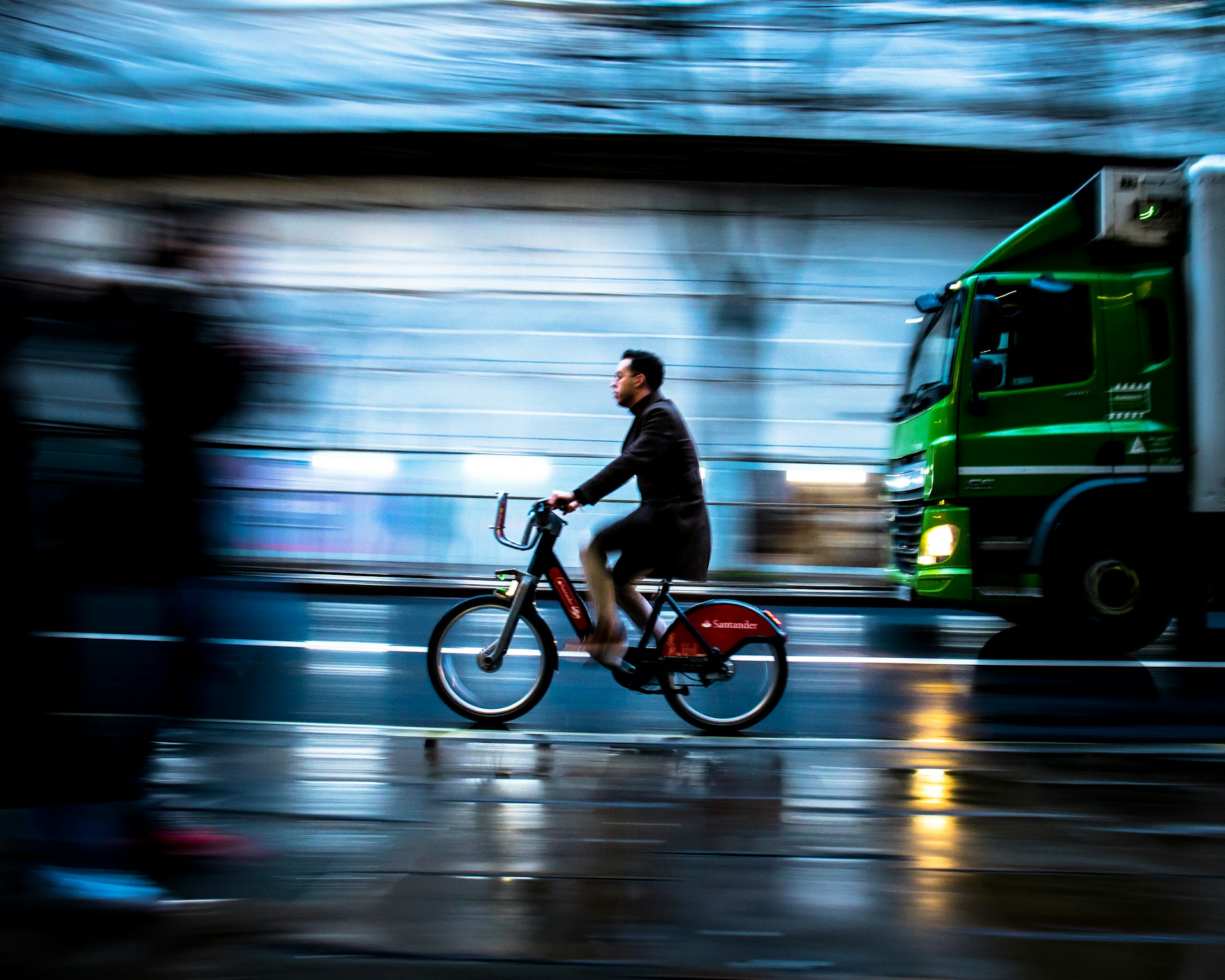 A motion-blurred urban scene featuring a person riding a red bike with a green truck passing by. The background is blurred, giving a sense of speed and movement. The wet pavement reflects city lights, enhancing the dynamic atmosphere.