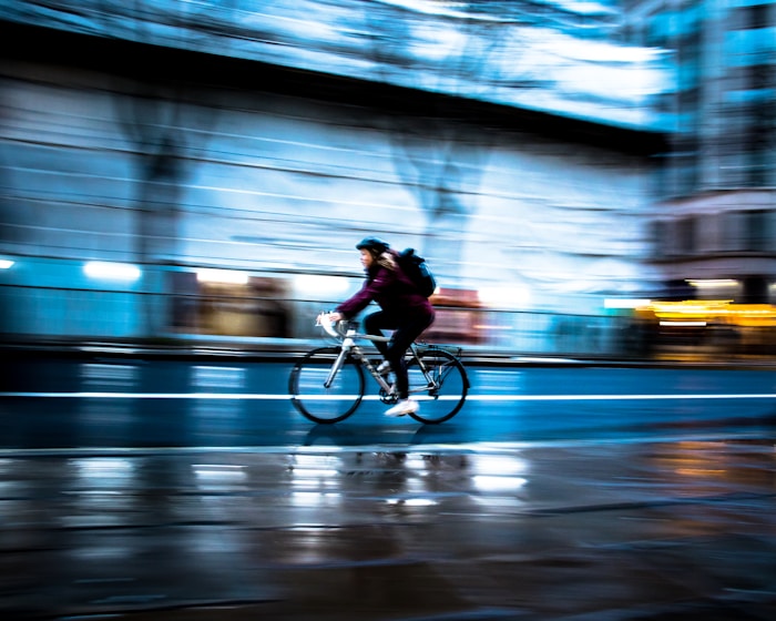 A cyclist is captured mid-motion on a bicycle, creating a dynamic blur effect with a clear sense of speed. The setting is an urban street with wet pavement, reflecting streetlights and buildings in the background. The scene conveys a sense of movement and urban energy.
