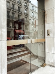 A glass entrance door with a metallic handle opens to reveal a staircase leading upwards. The building's facade reflects in the glass, showing intricate window designs and balconies. Nearby, a keypad is mounted on the wall beside the entrance.