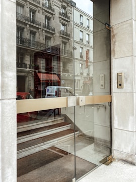 A glass entrance door with a metallic handle opens to reveal a staircase leading upwards. The building's facade reflects in the glass, showing intricate window designs and balconies. Nearby, a keypad is mounted on the wall beside the entrance.