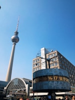 A tall television tower stands prominently against a clear blue sky, with a large, round world clock in the foreground displaying various city times. The surrounding area includes modern architecture with glass and steel elements.
