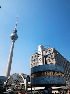 A tall television tower stands prominently against a clear blue sky, with a large, round world clock in the foreground displaying various city times. The surrounding area includes modern architecture with glass and steel elements.