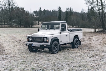 A white Land Rover Defender is parked on a frosty, grassy field. The weather is overcast, and the surrounding area contains some sparse trees and a distant treeline. The vehicle appears rugged and well-suited for off-road conditions.