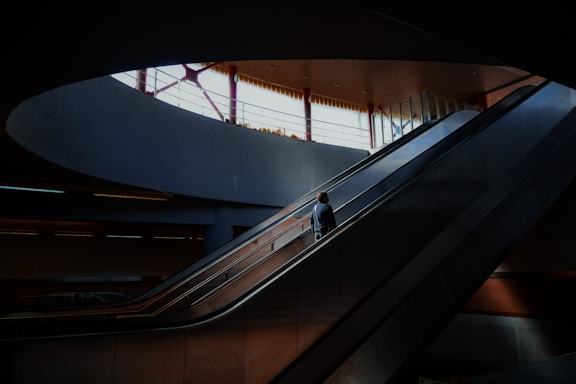 A solitary figure ascends an escalator in a spacious indoor setting, illuminated by natural light streaming through large windows above. The architecture features clean lines and a modern design, with a muted color palette creating a serene atmosphere.