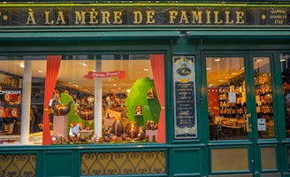 A storefront with a vintage design features green wooden frames and gold lettering reading 'A La Mère de Famille.' The display window is decorated with various festive items, including large chocolate eggs and colorful ornaments. Inside, the shop is filled with shelves that appear to hold a range of confectionery.