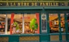 A storefront with a vintage design features green wooden frames and gold lettering reading 'A La M&egrave;re de Famille.' The display window is decorated with various festive items, including large chocolate eggs and colorful ornaments. Inside, the shop is filled with shelves that appear to hold a range of confectionery.