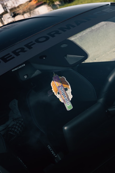 A car interior is shown with a focus on the sticker attached to the windshield. The sticker features colorful artwork and the word 'mentos' is prominently displayed. The interior includes a Recaro seat, and the word 'PERFORMANCE' is visible on the windshield's sunstrip.