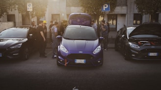 A group of people gathers around three parked cars on a street, with the central car in a vibrant blue color and its trunk open. The surrounding people appear engaged in conversation, possibly discussing the vehicles. A 'P' parking sign is visible above, indicating legal parking.