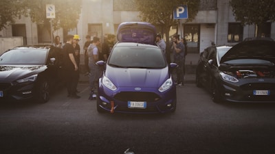 A group of people gathers around three parked cars on a street, with the central car in a vibrant blue color and its trunk open. The surrounding people appear engaged in conversation, possibly discussing the vehicles. A 'P' parking sign is visible above, indicating legal parking.
