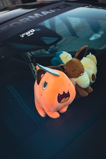 Three plush toys representing characters are placed on the dashboard of a car. They include an orange dinosaur-like creature, a brown fox-like creature, and a blue and yellow bear-like creature. The toys are positioned beside a car sticker, and the lighting suggests an outdoor setting.