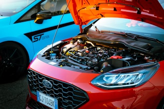A bright red car with an open hood displaying its engine. The front grille features the Ford logo, and the headlight is visible. In the background, a blue car marked with 'ST Edition' on the door is partially visible. The scene is well-lit, suggesting it takes place outdoors during the day.