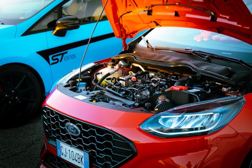 A bright red car with an open hood displaying its engine. The front grille features the Ford logo, and the headlight is visible. In the background, a blue car marked with 'ST Edition' on the door is partially visible. The scene is well-lit, suggesting it takes place outdoors during the day.