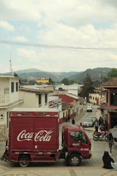 A red Coca-Cola truck is parked on a sloped street in a town with colonial architecture. The street is lined with buildings with red tile roofs and colorful facades. People are walking and interacting in the vicinity. In the background, mountains and a visible church add to the scenic view.