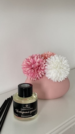 A pink ceramic vase holds three artificial flowers in shades of white and pink, placed on a white surface. Beside it, there is a small bottle of fragrance with black-reed diffusers. The bottle has a black label with white and gold text.